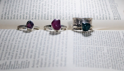 Green-blue stone spinner ring with a wide hammered silver band, pictured beside two purple and pink stone rings on an open book.