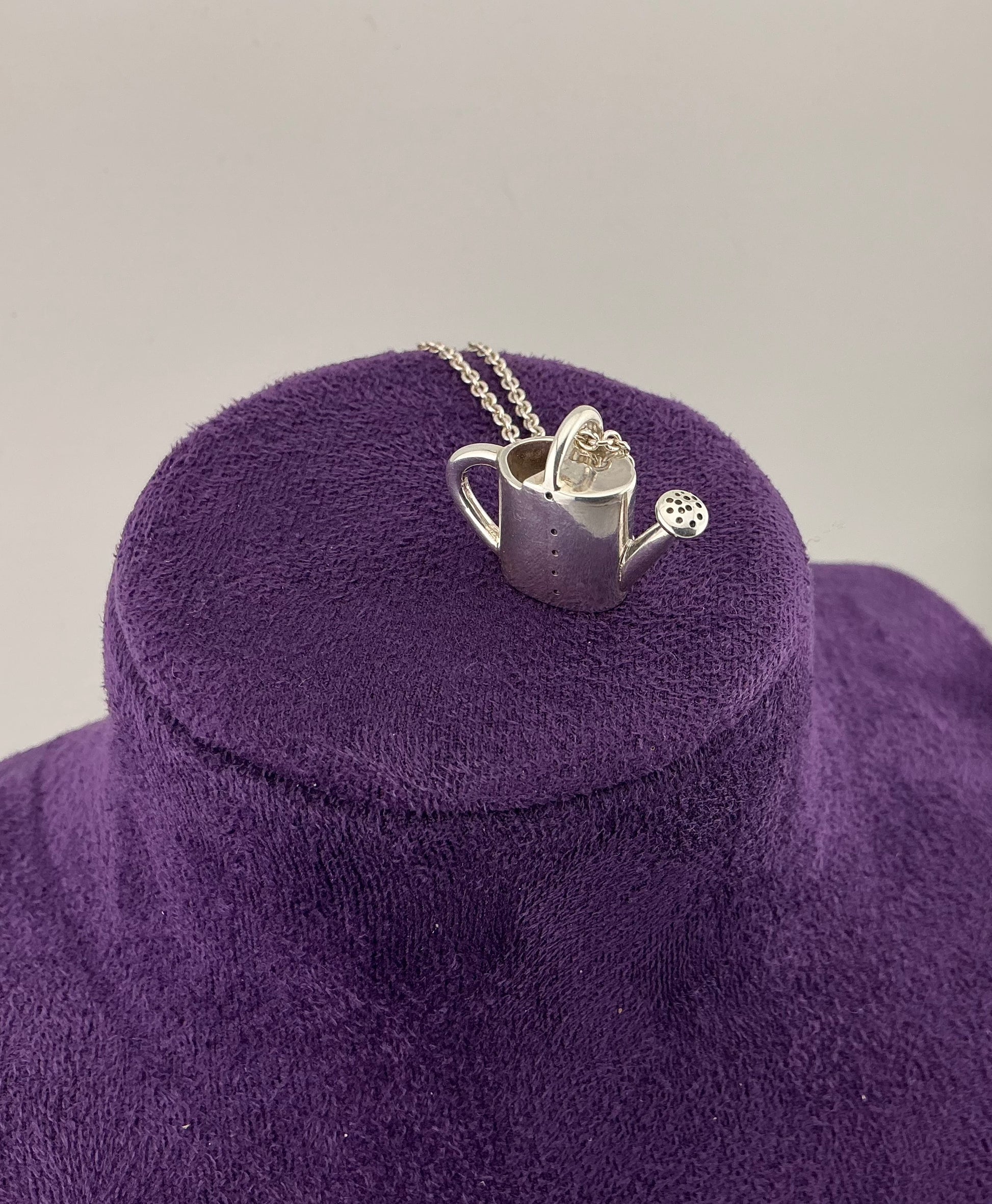 Silver watering can pendant on a purple cushion