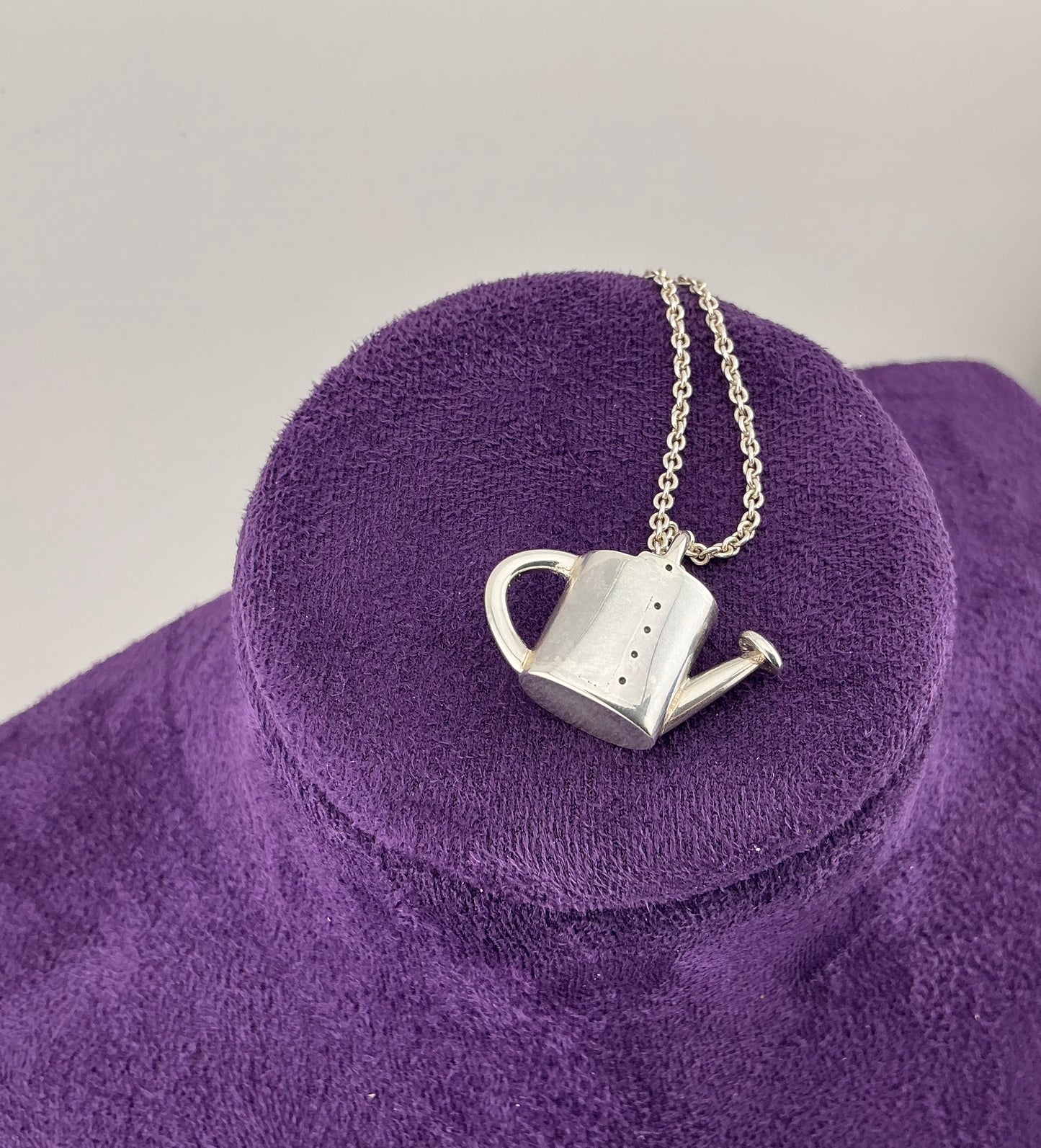 Silver watering can pendant necklace on a purple stand with a gray background