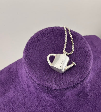 Silver watering can pendant necklace on a purple stand with a gray background