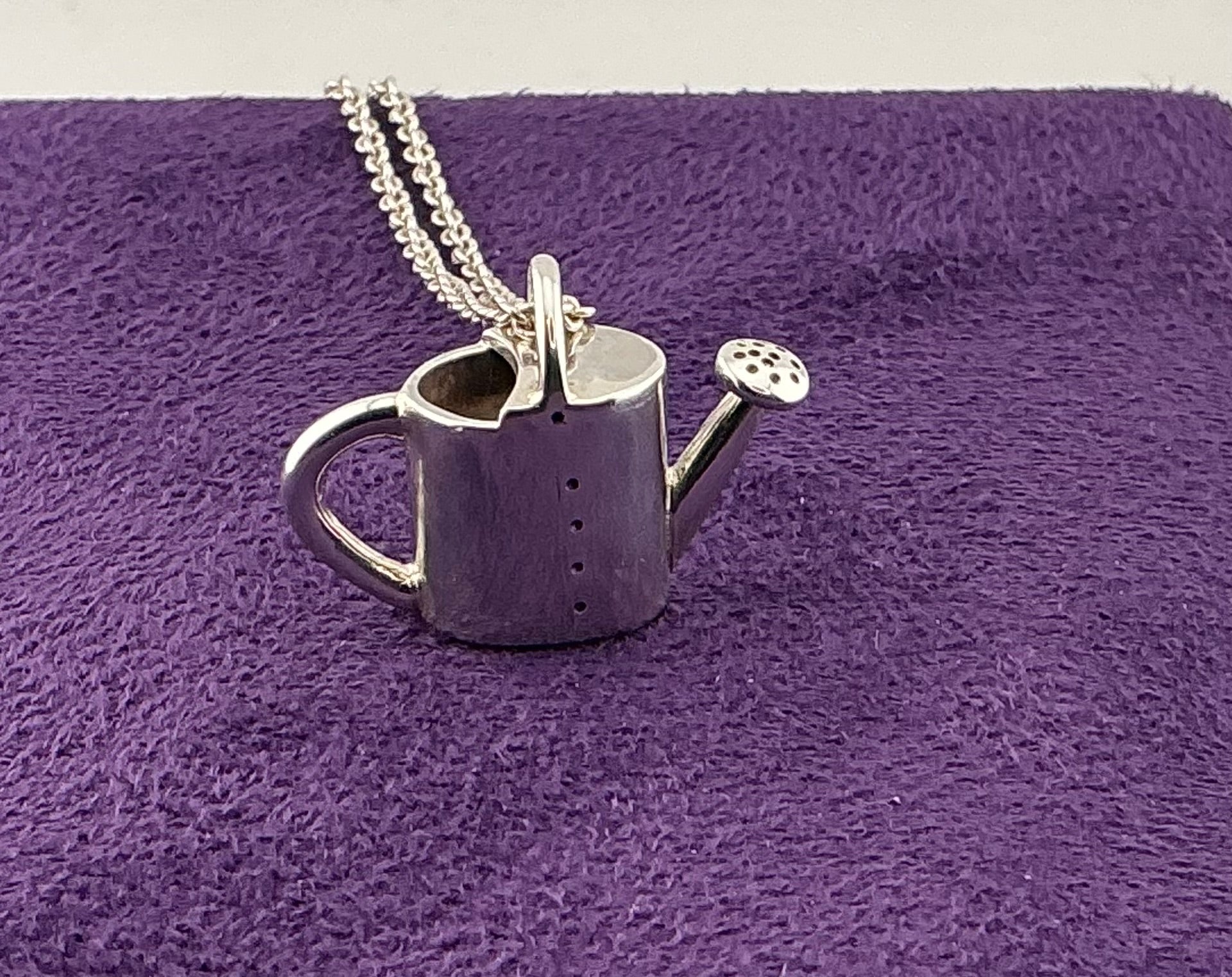 Silver watering can pendant on a chain against a purple textured background