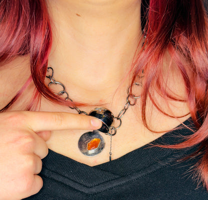 Person wearing a unique necklace with a central orange stone and chain design, pointing at it.