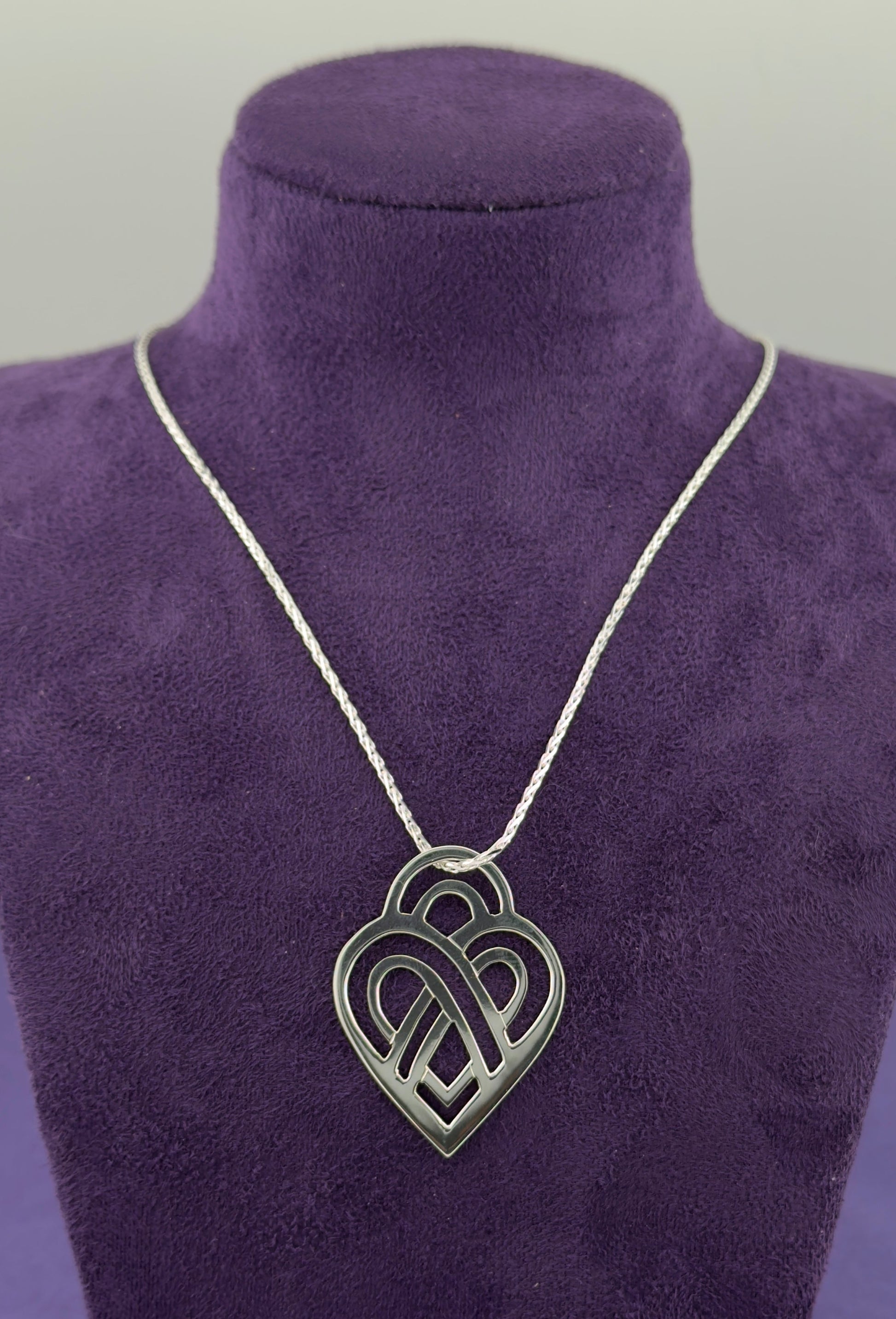 Silver necklace with a heart-shaped pendant on a purple velvet stand.