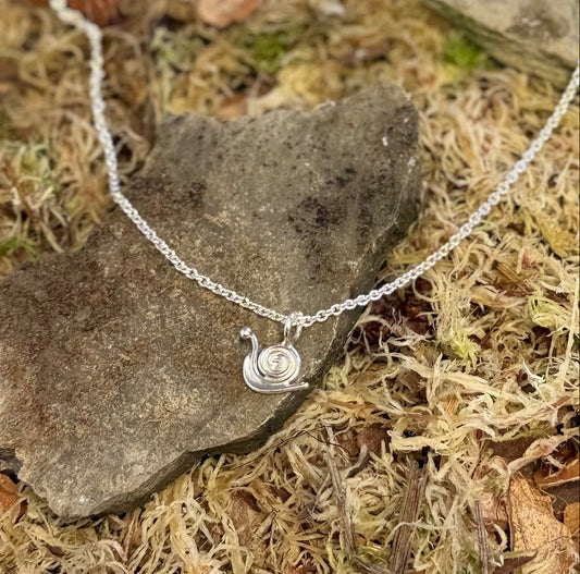 Silver necklace with a snail pendant on a stone surface with moss and small stones.