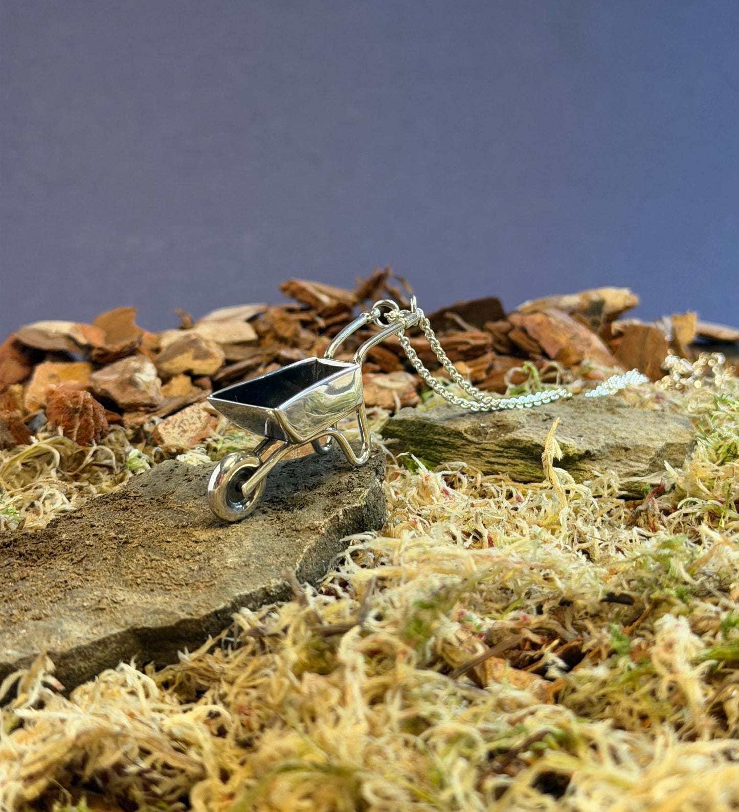 Silver necklace with a wheelbarrow pendant on a rocky and mossy surface
