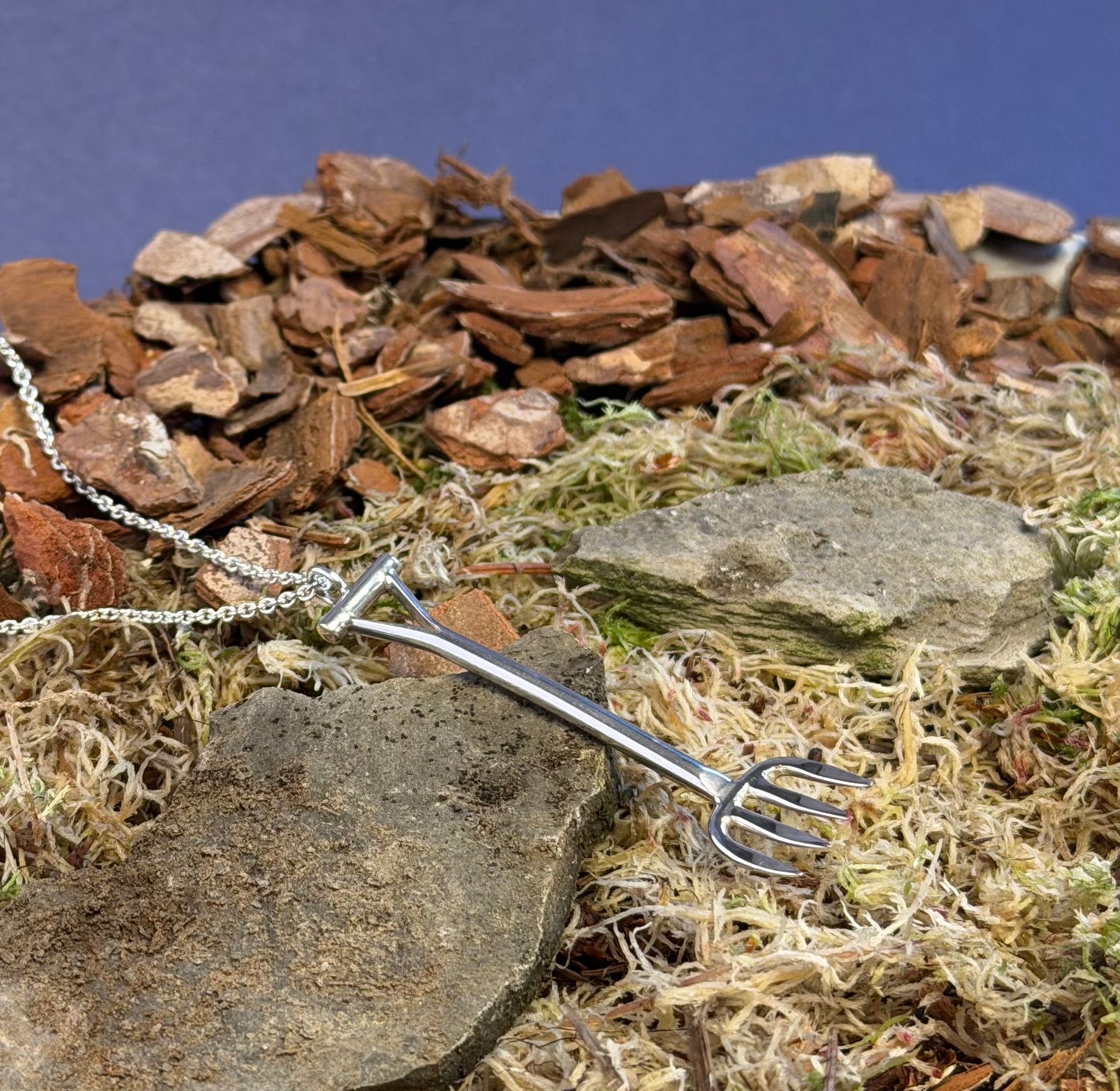 Silver garden fork-shaped pendant on a chain against a natural background of rocks and moss.