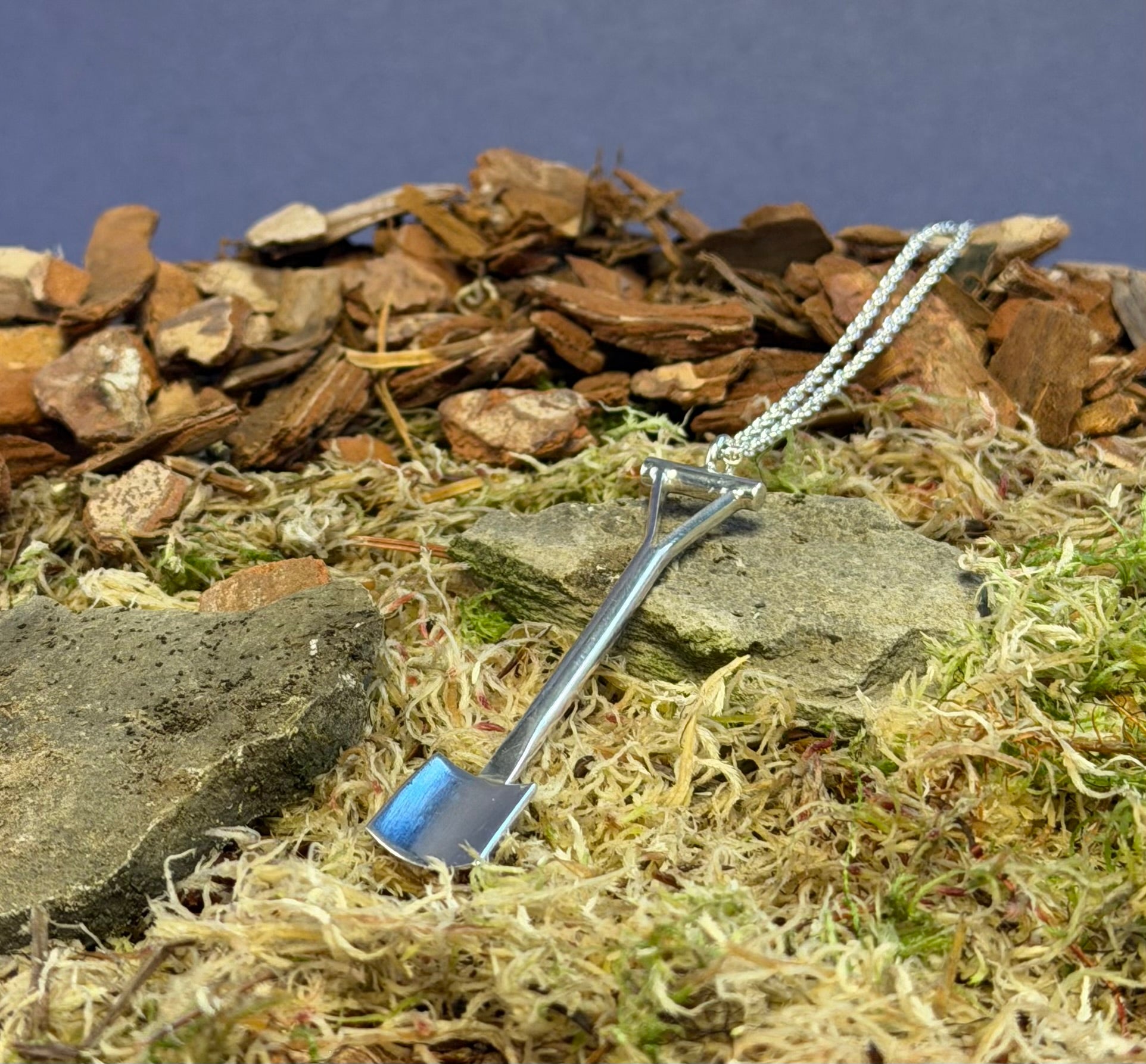 Silver spade pendant on a chain against a natural background of rocks and moss.