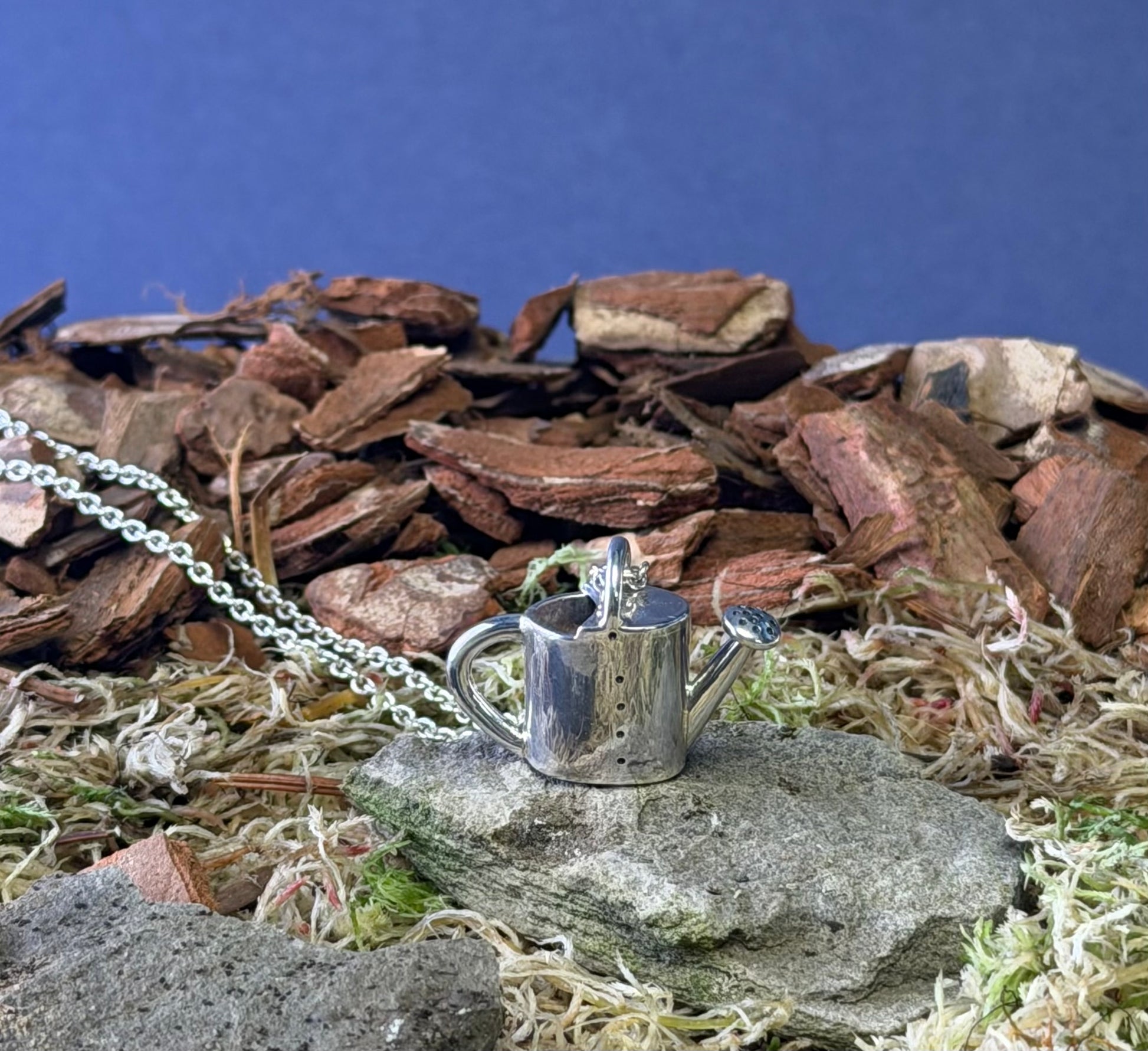 Silver watering can pendant on a chain against a natural background with stones and grass.