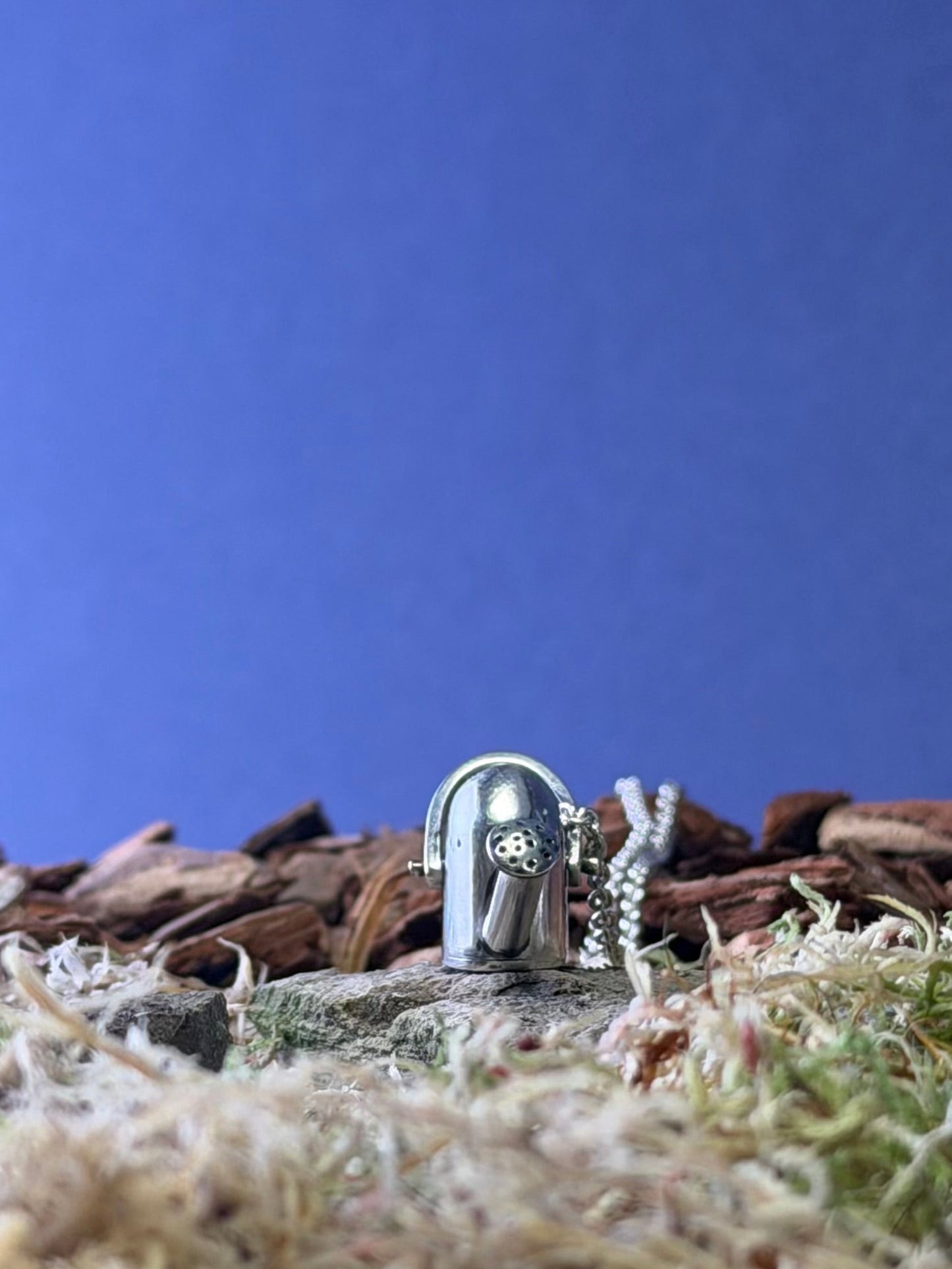 Small watering can pendant on chain on a miniature landscape with a blue sky background