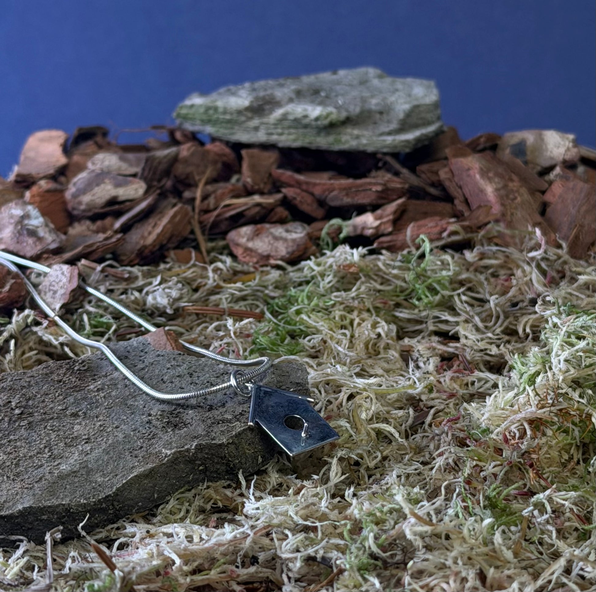 Close-up of a miniature birdhouse necklace on a  landscape with rocks and moss on a blue background