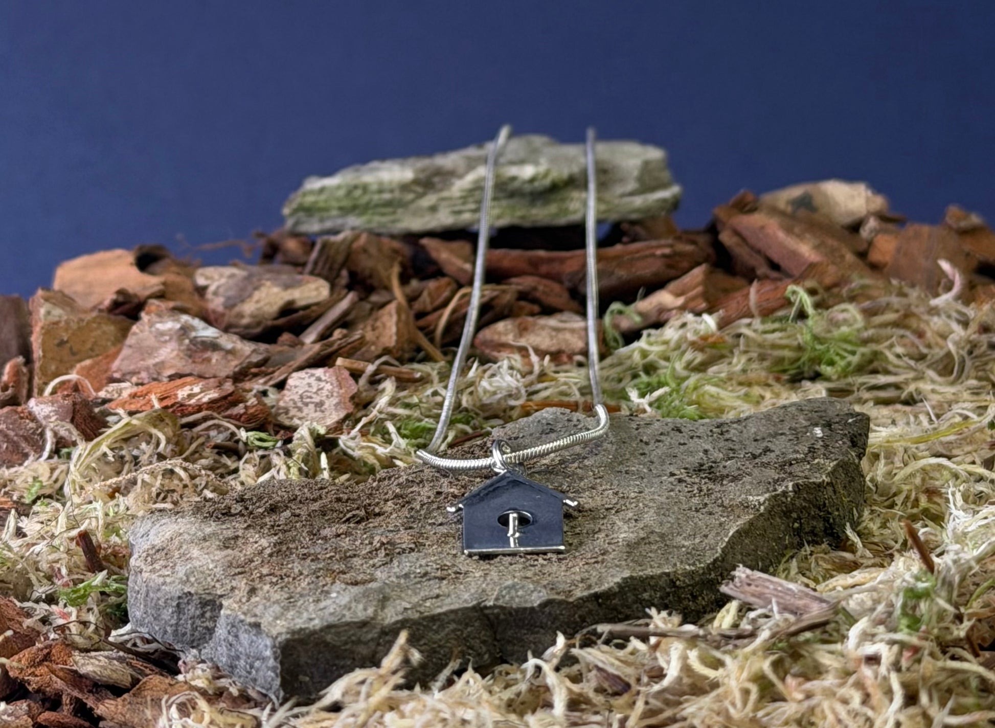 Small birdhouse shaped pendant on a chain against a natural background with stones and plants.