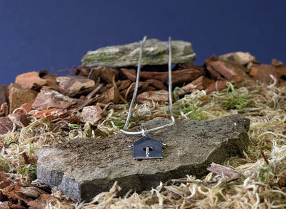 Small birdhouse shaped pendant on a chain against a natural background with stones and plants.