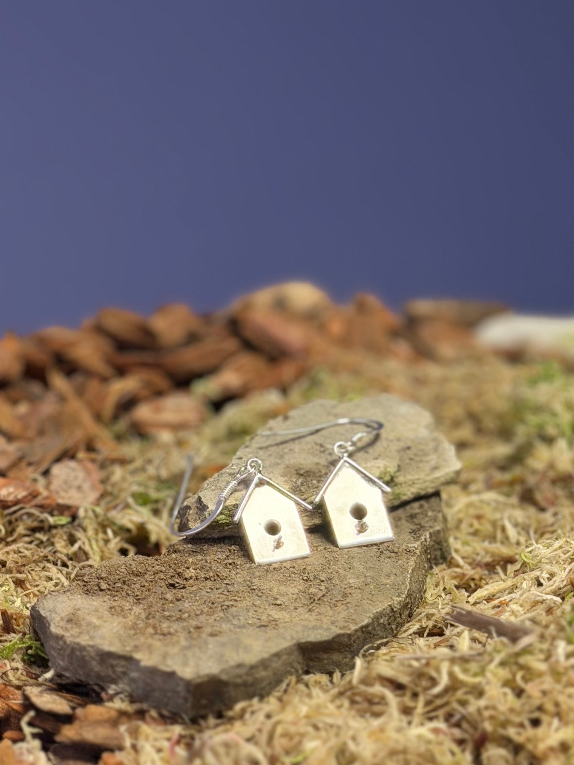 Small birdhouse shaped earrings on a stone slab with moss and small stones in the background