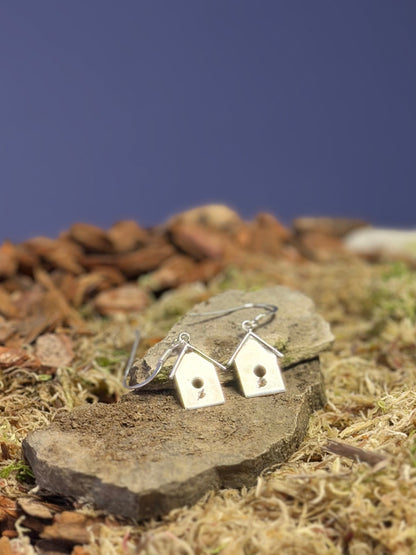 Small birdhouse shaped earrings on a stone slab with moss and small stones in the background