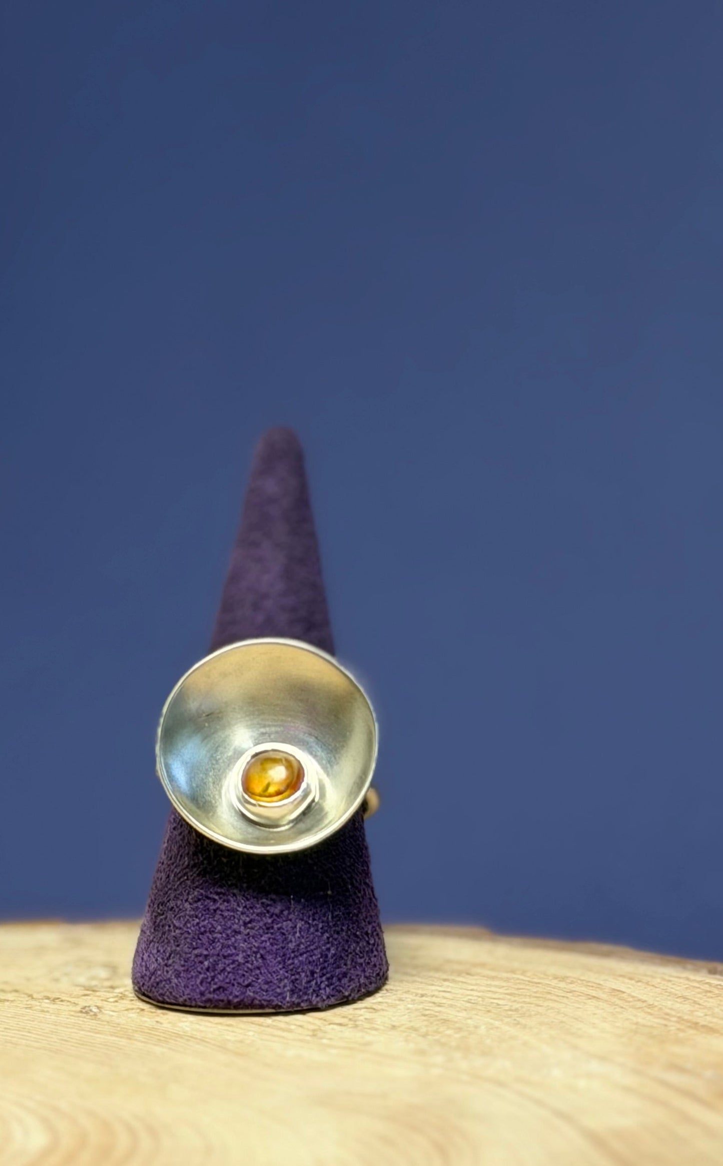Silver ring set with an amber stone, with a purple cone on a wooden surface against a blue background