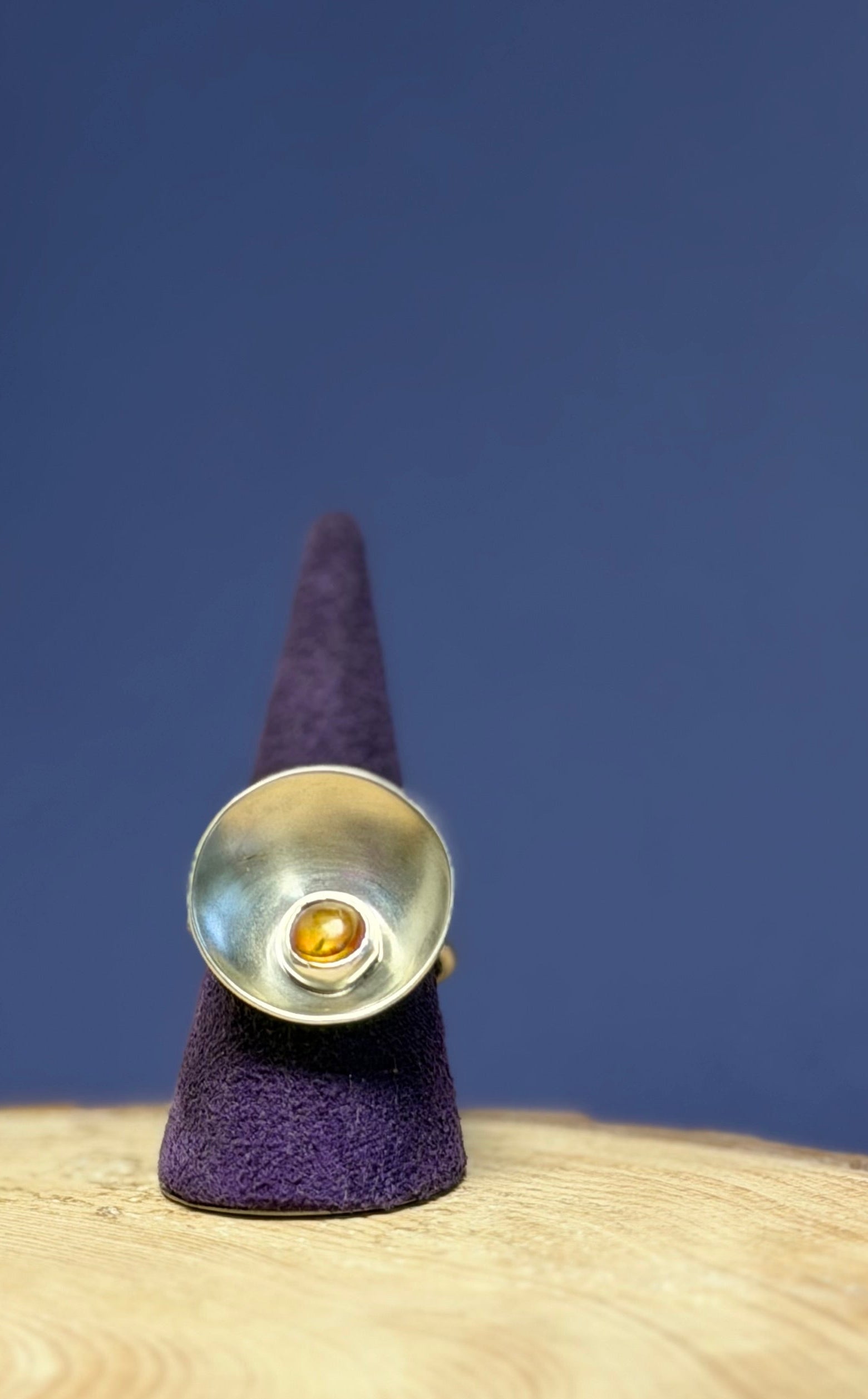 Silver ring set with an amber stone, with a purple cone on a wooden surface against a blue background