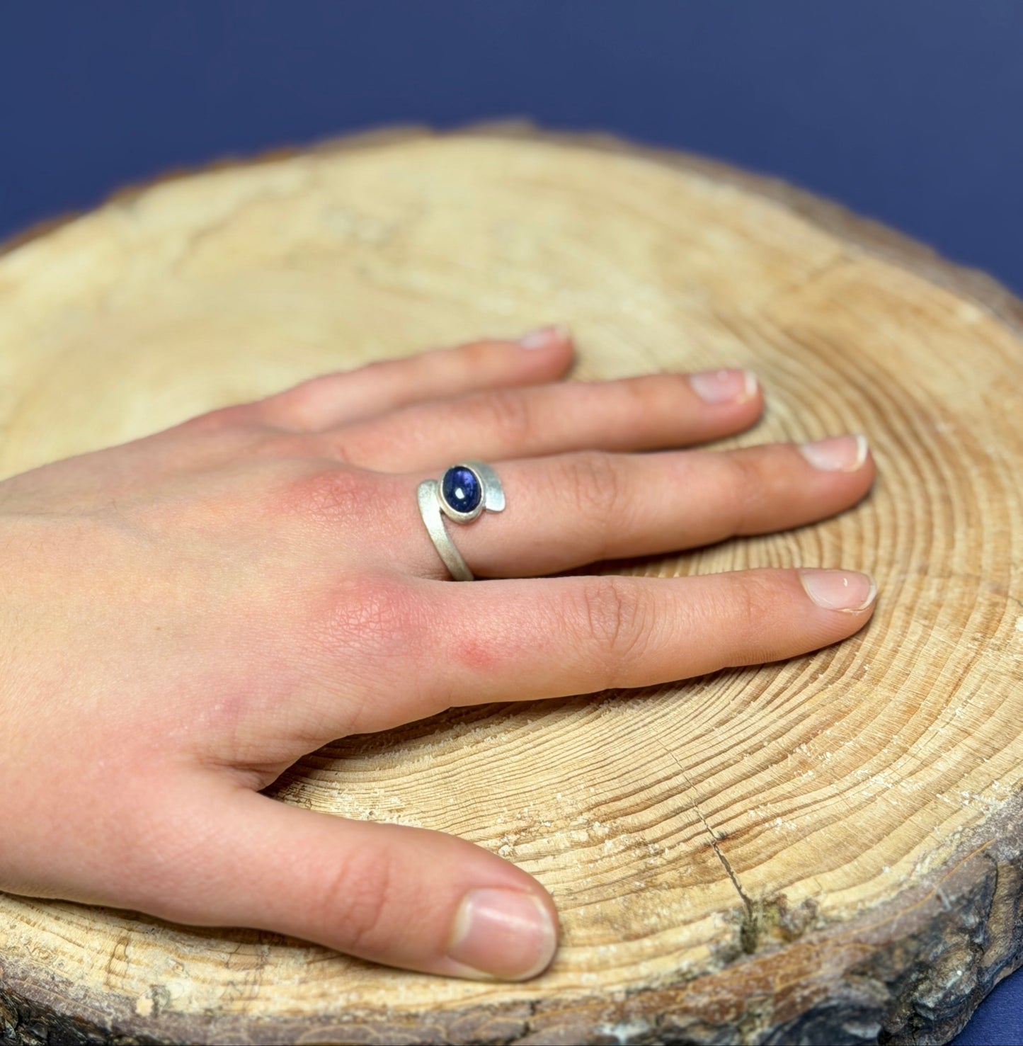 Hand wearing a silver ring with a blue purple gemstone on a wooden surface with a blue background