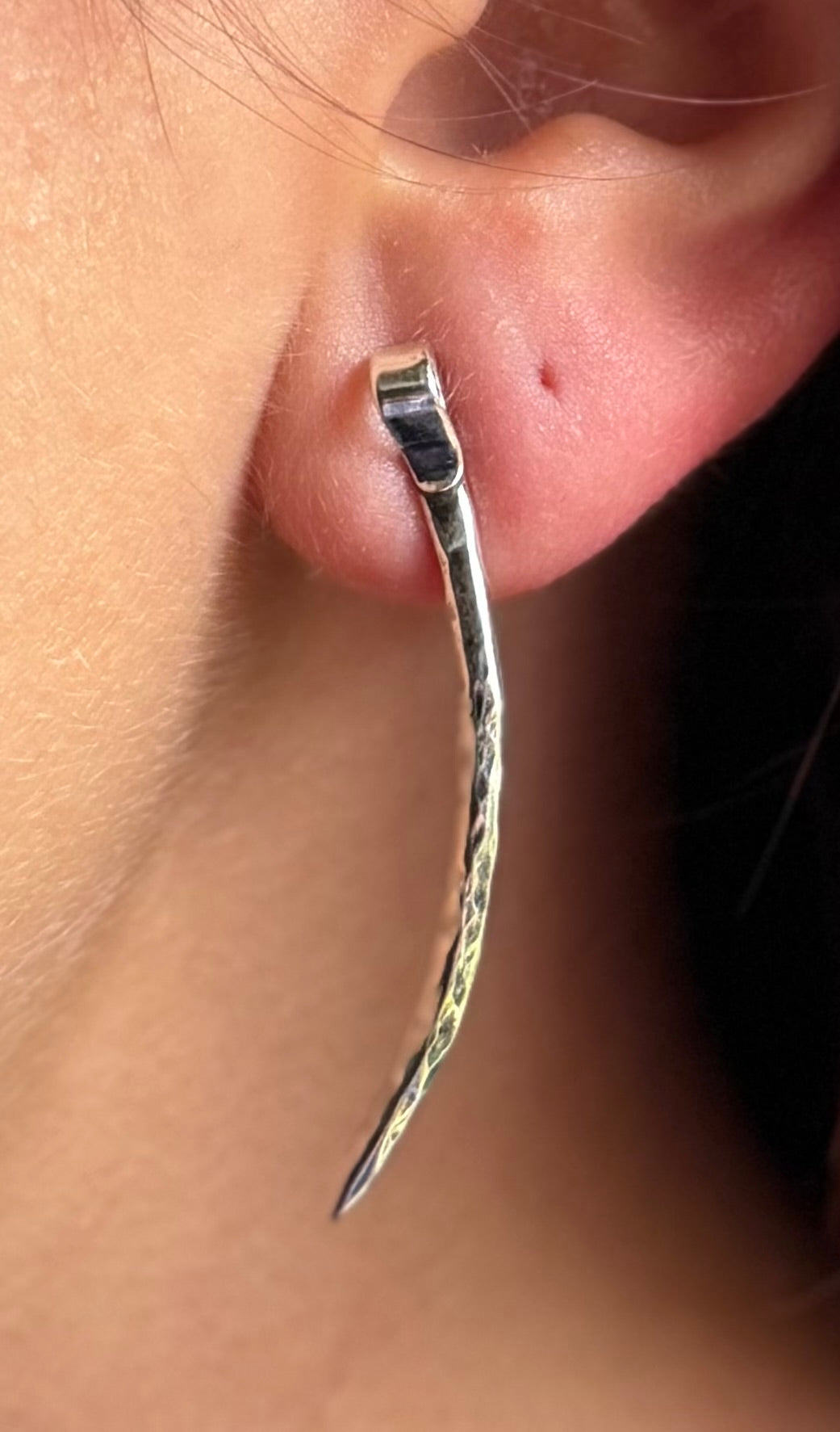 Close-up of a long forged silver spike stud worn on an earlobe, showing its textured surface and curved point.