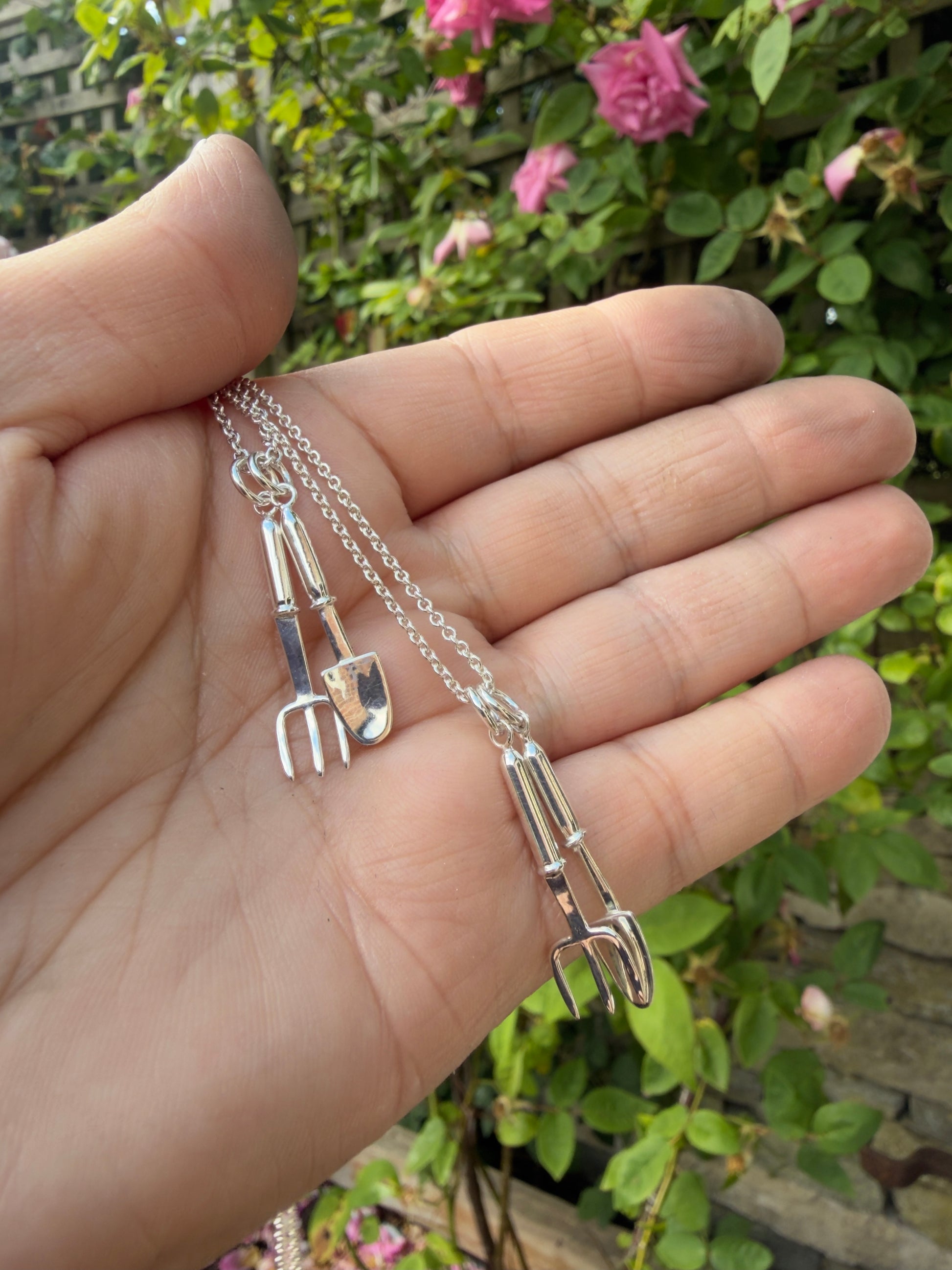 Silver miniature trowel and hand fork on belcher chain, in hand with rose bushes in the background.