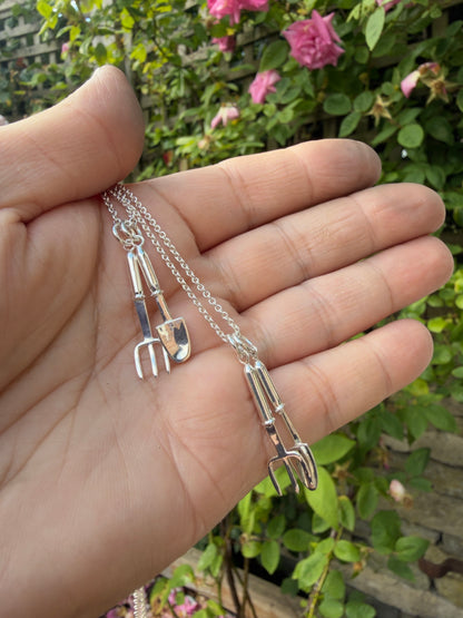 Silver miniature trowel and hand fork on belcher chain, in hand with rose bushes in the background.