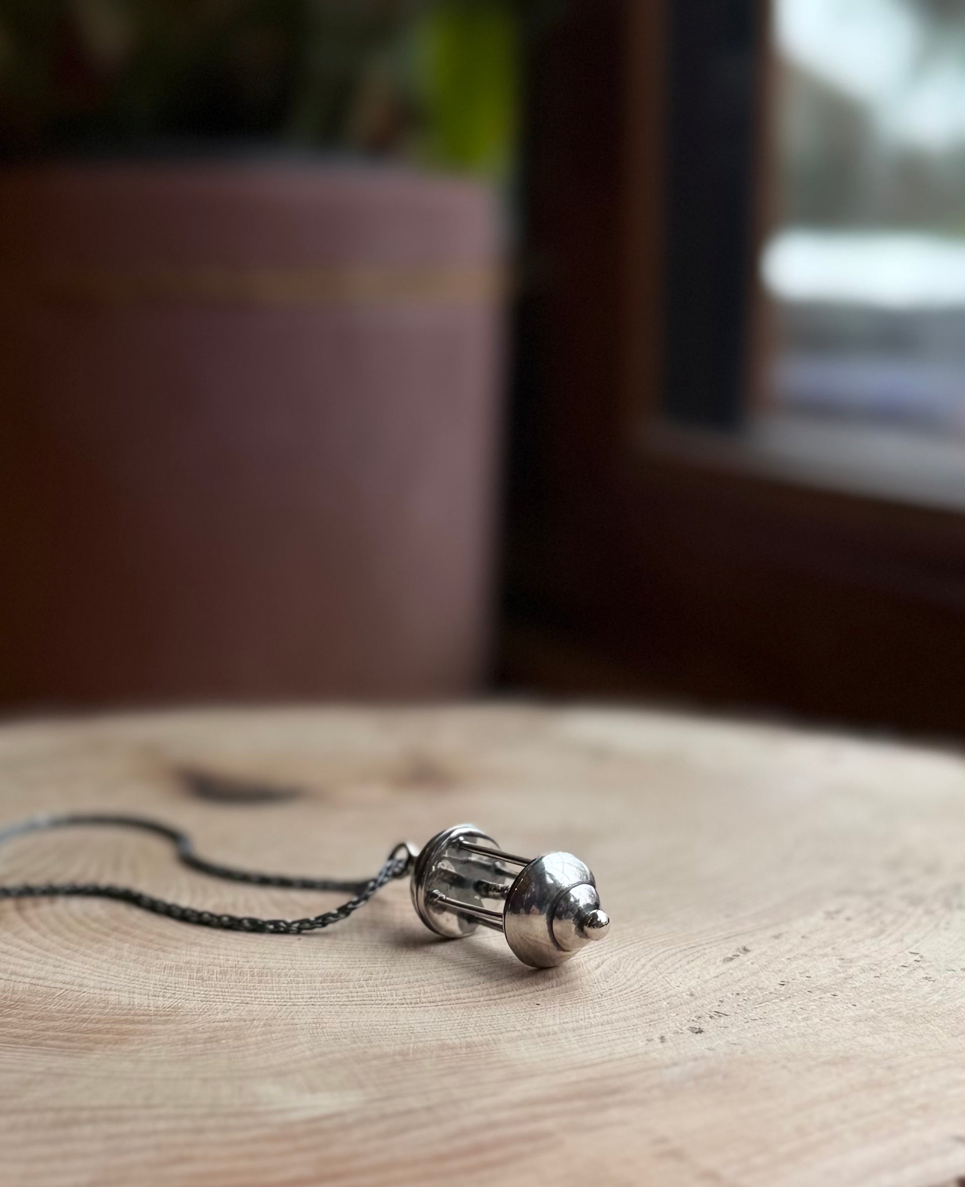 Silver lantern pendant, resting on wood, with a plant pot in the background