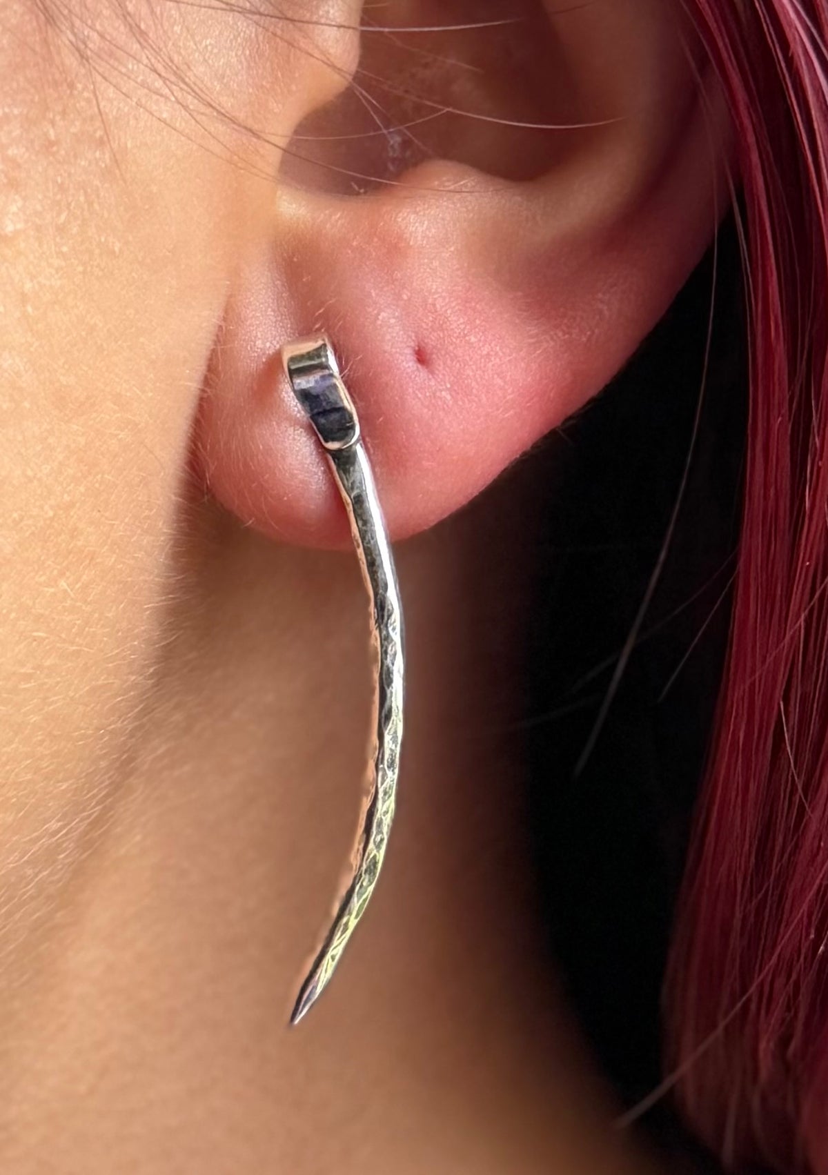 Long forged silver spike stud worn beside red hair, showing its elongated curve and tapered point.