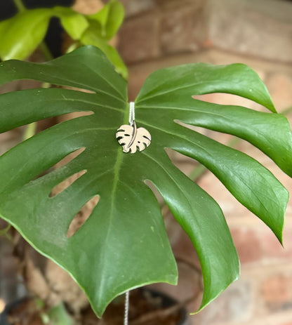 Monstera leaf pendant made in silver, draped over a monstera leaf with a brick background.