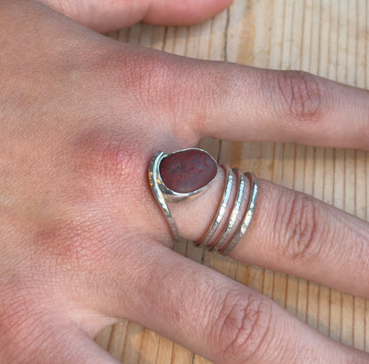 Reddish-brown beach pebble spiral ring worn on a finger, showing the silver setting and hammered band wrapping around the finger.