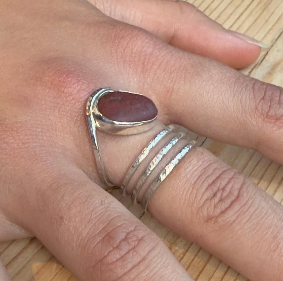 Angled view of a reddish-brown beach pebble ring worn on a hand, showing the raised stone setting and open spiral band.