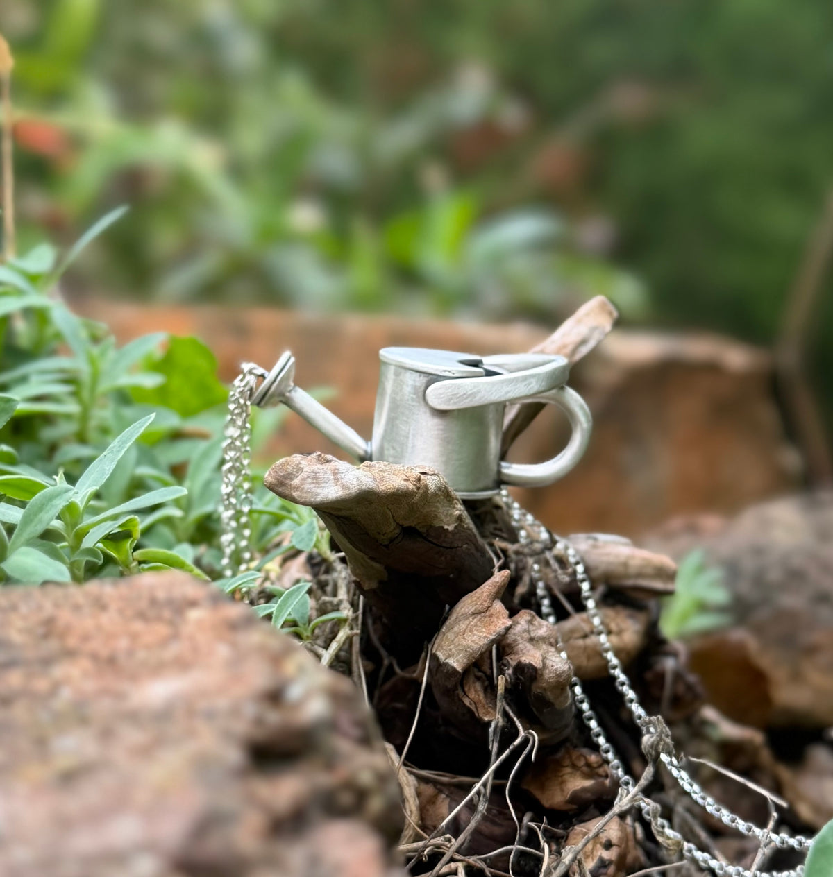 Side view of a silver watering can pendant resting on wood among green plants, with chain strands falling from its spout to symbolise water.