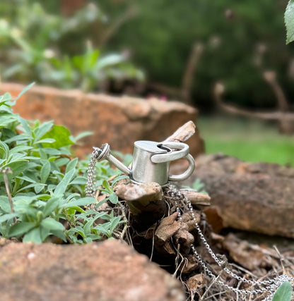 Silver watering can pendant on weathered wood, showing its curved handles, long spout and cascading chain details.