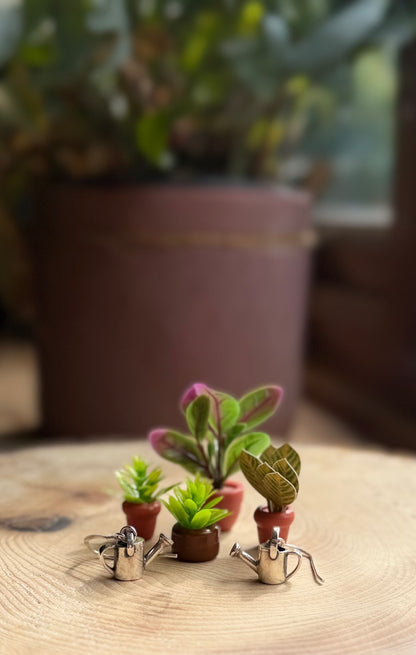 Silver watering can drop earrings, next to fake miniature plant pots and flowers
