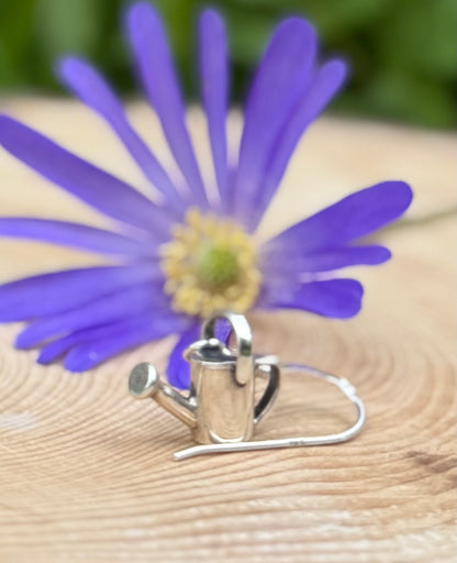 Silver watering can drop earrings, next to flowers