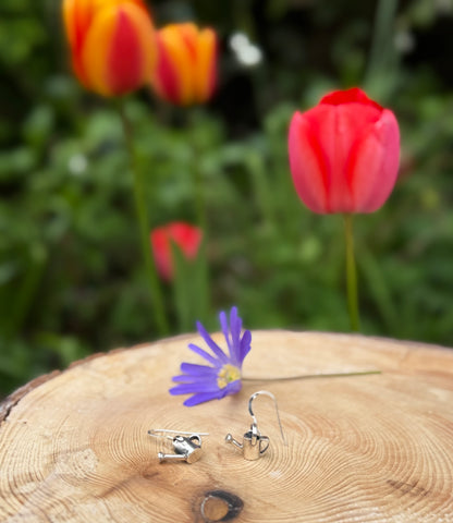 Silver watering can drop earrings, next to flowers