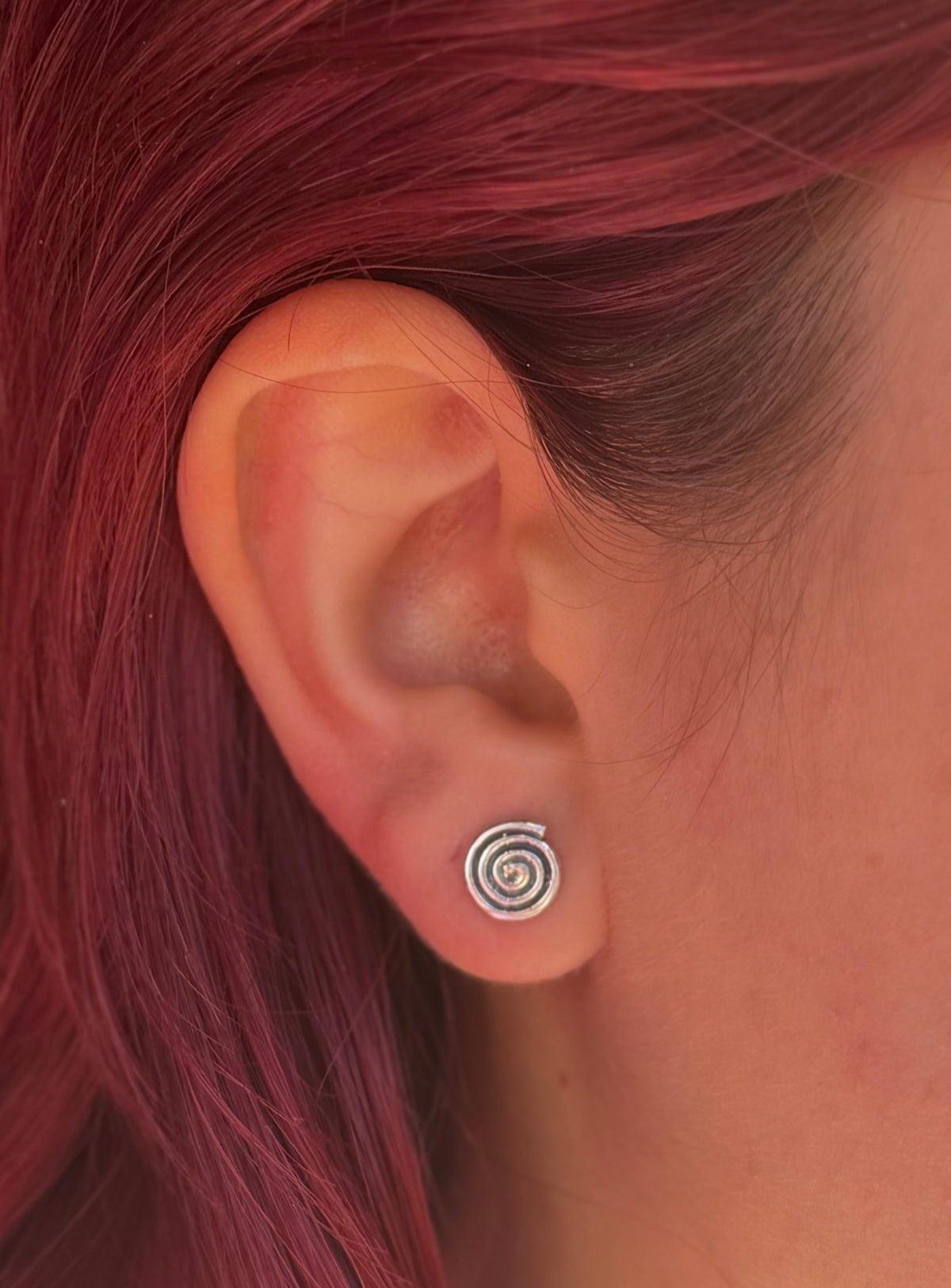 Colour close-up of an oxidised silver spiral stud earring worn beside red hair, showing its coiled design.