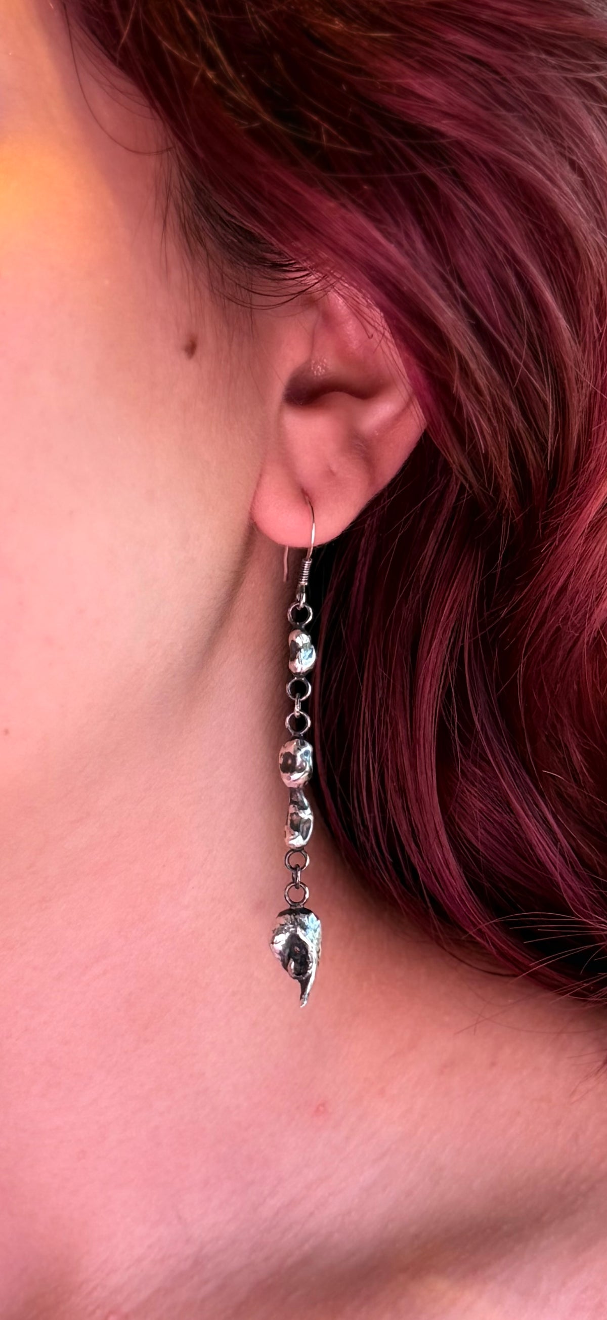 Colour close-up of a triple silver nugget drop earring worn beside red hair, showing its sculptural shapes and reflective surfaces.