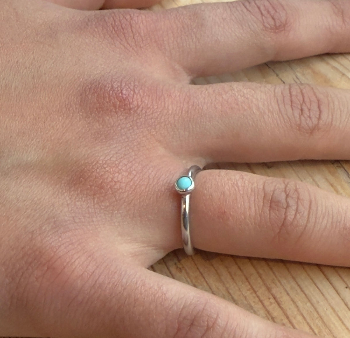 Silver ring with a small round turquoise cabochon, worn alone on a finger resting on a wooden surface.