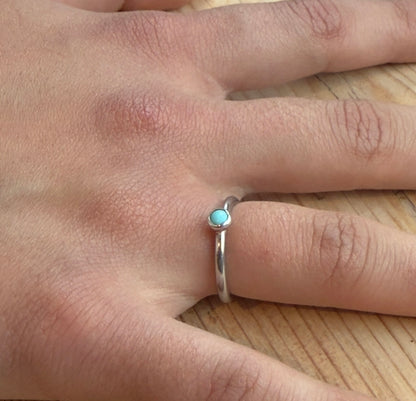 Silver ring with a small round turquoise cabochon, worn alone on a finger resting on a wooden surface.