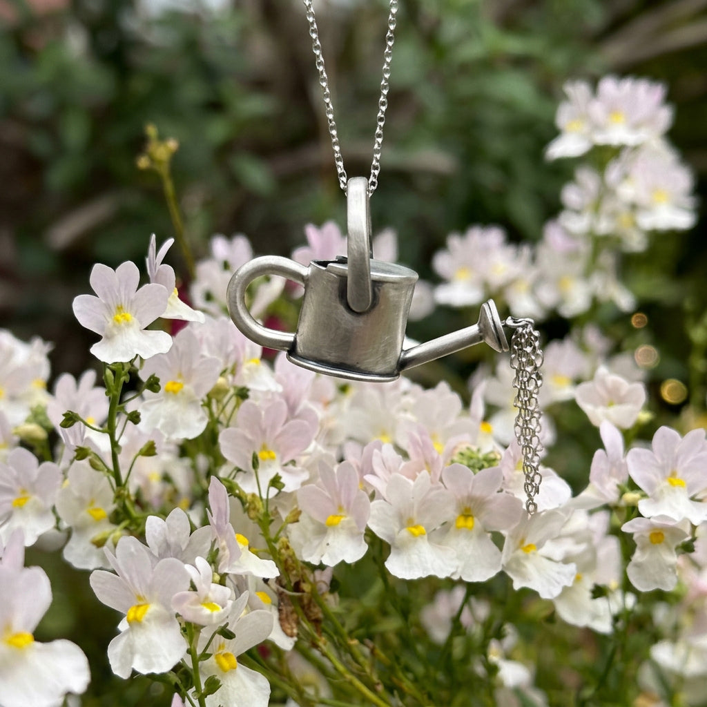 Silver watering can pendant suspended above pale flowers, with delicate chain strands pouring from its spout.