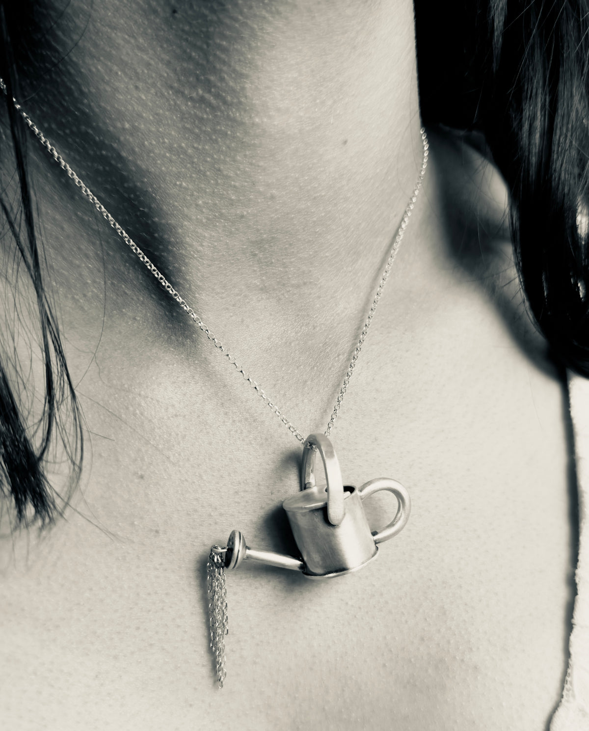Silver watering can pendant worn on a necklace, with delicate chain strands hanging from the spout like pouring water. Black and white image.