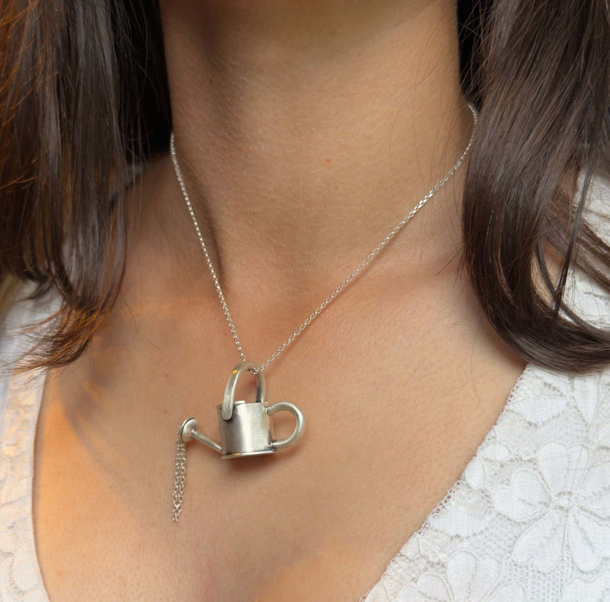 Silver watering can pendant worn on a necklace, with delicate chain strands hanging from the spout like pouring water.