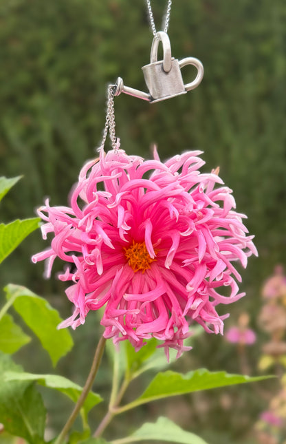 Silver watering can pendant hanging above a pink flower, with dangling chains creating the effect of pouring water.