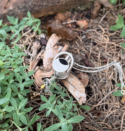 Top view of a silver watering can pendant on wood among green leaves, showing its open top, handle and detailed spout.