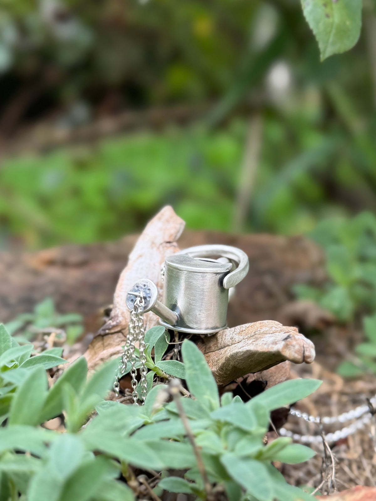 Silver watering can pendant nestled among garden plants and wood, with dangling chains representing pouring water.