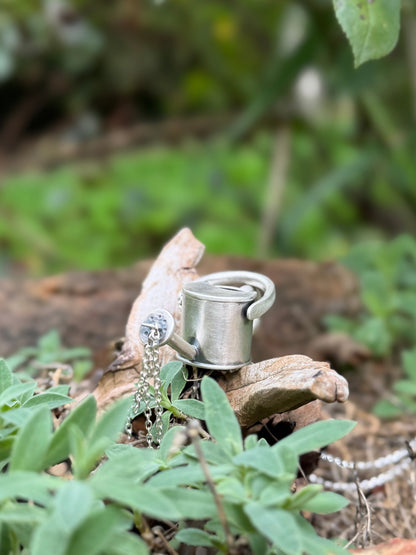 Silver watering can pendant nestled among garden plants and wood, with dangling chains representing pouring water.