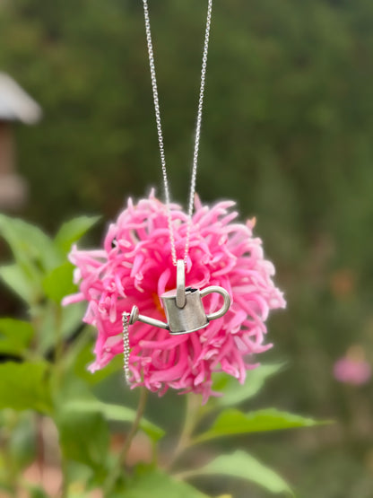 Watering can pendant on a silver-coloured necklace chain in front of a pink flower, showing its miniature garden design.