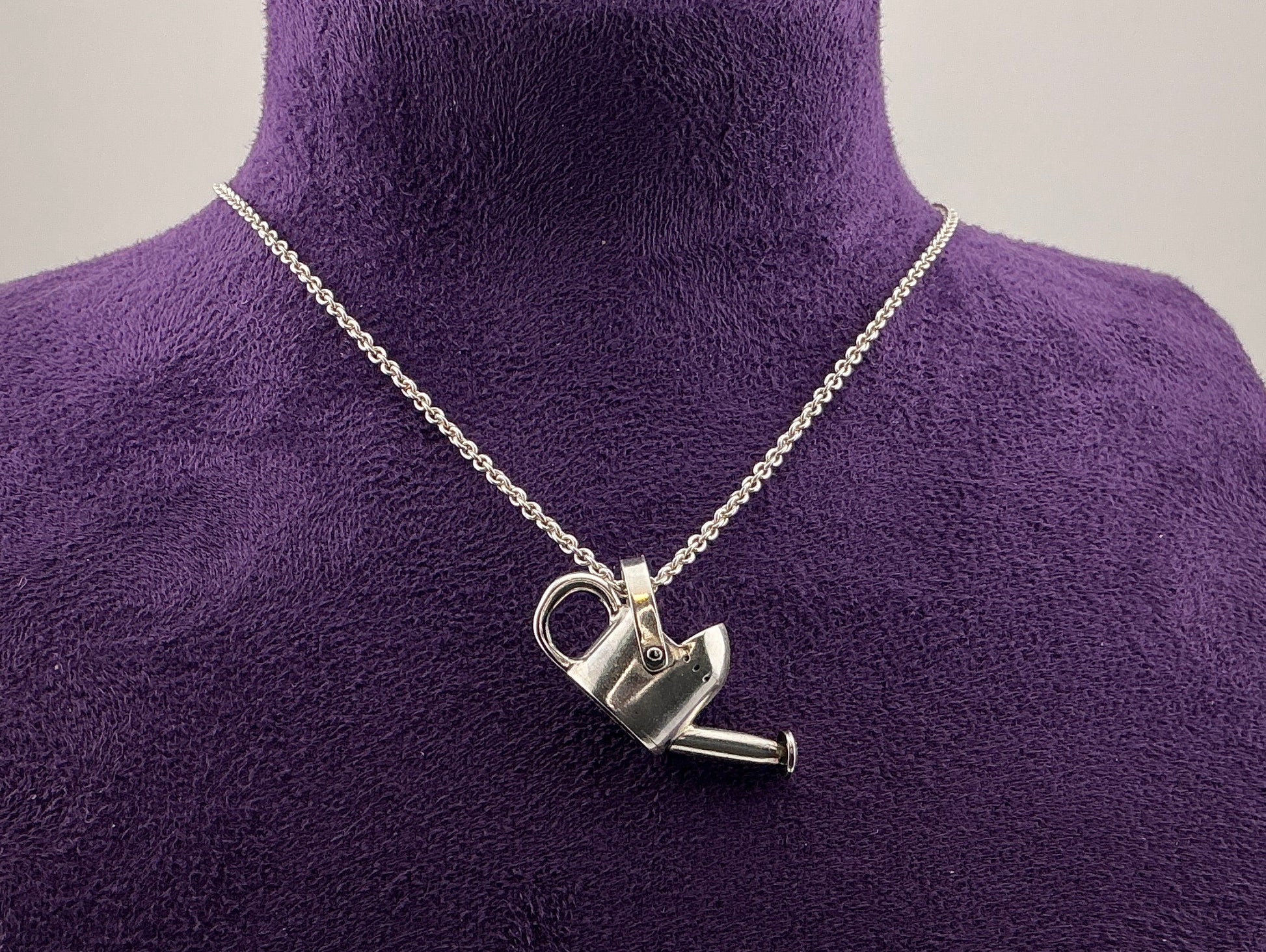 Silver necklace with a watering can pendant on a purple mannequin.